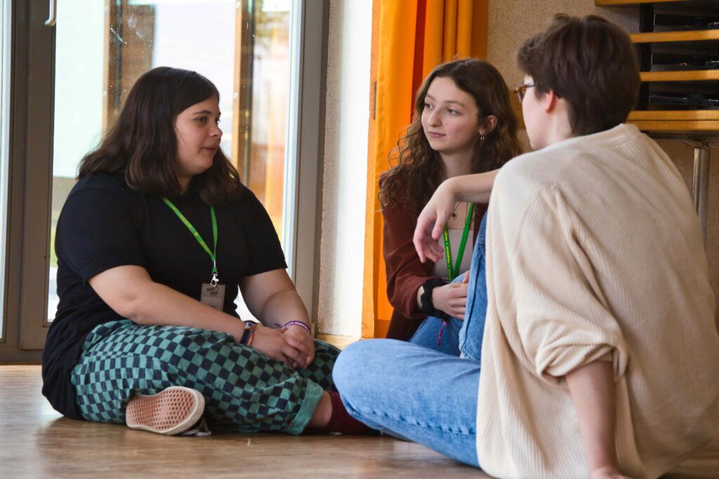 Drei junge Menschen sitzen vor einem Fenster auf dem Boden und unterhalten sich.