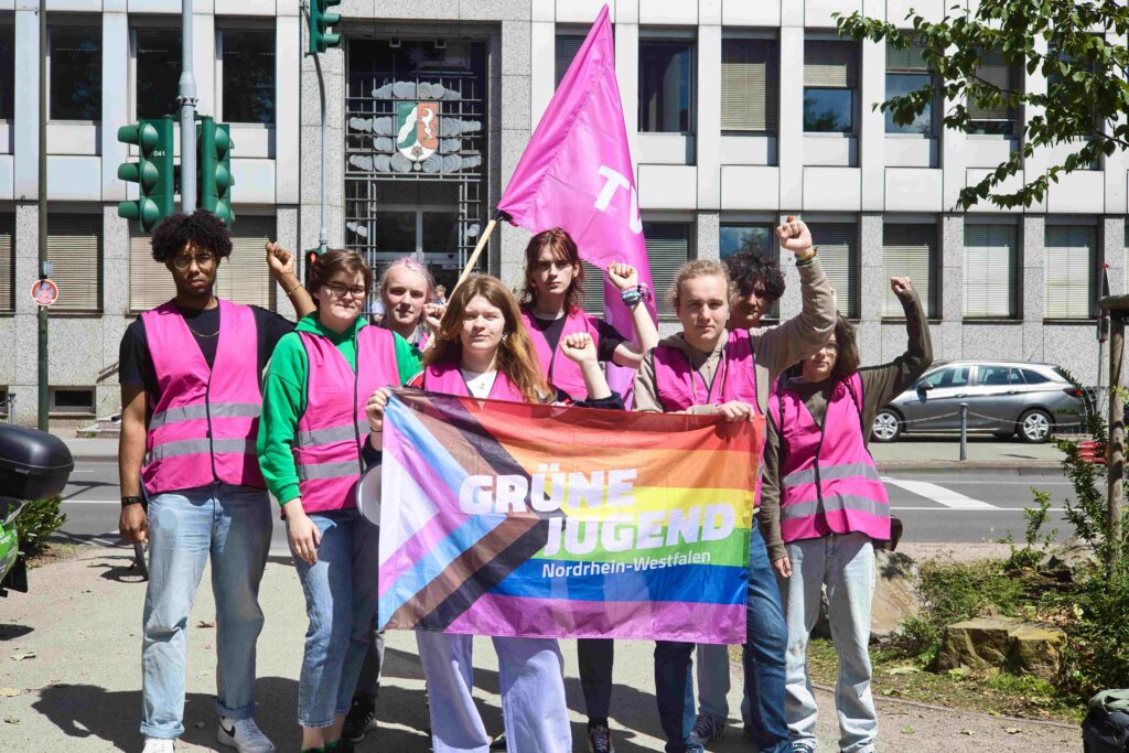 Acht junge Menschen in pinken Warnwesten halten die Linke Faust in die Luft und halten eine Fahne mit der Aufschrift: "Grüne Jugend Nordrhein-Westfalen"