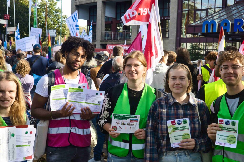 Fünf junge Menschen stehen in einer Demonstration und halten Flyer und Unterschriftenlisten der Wir-fahren-zusammen-Kampgne hoch.